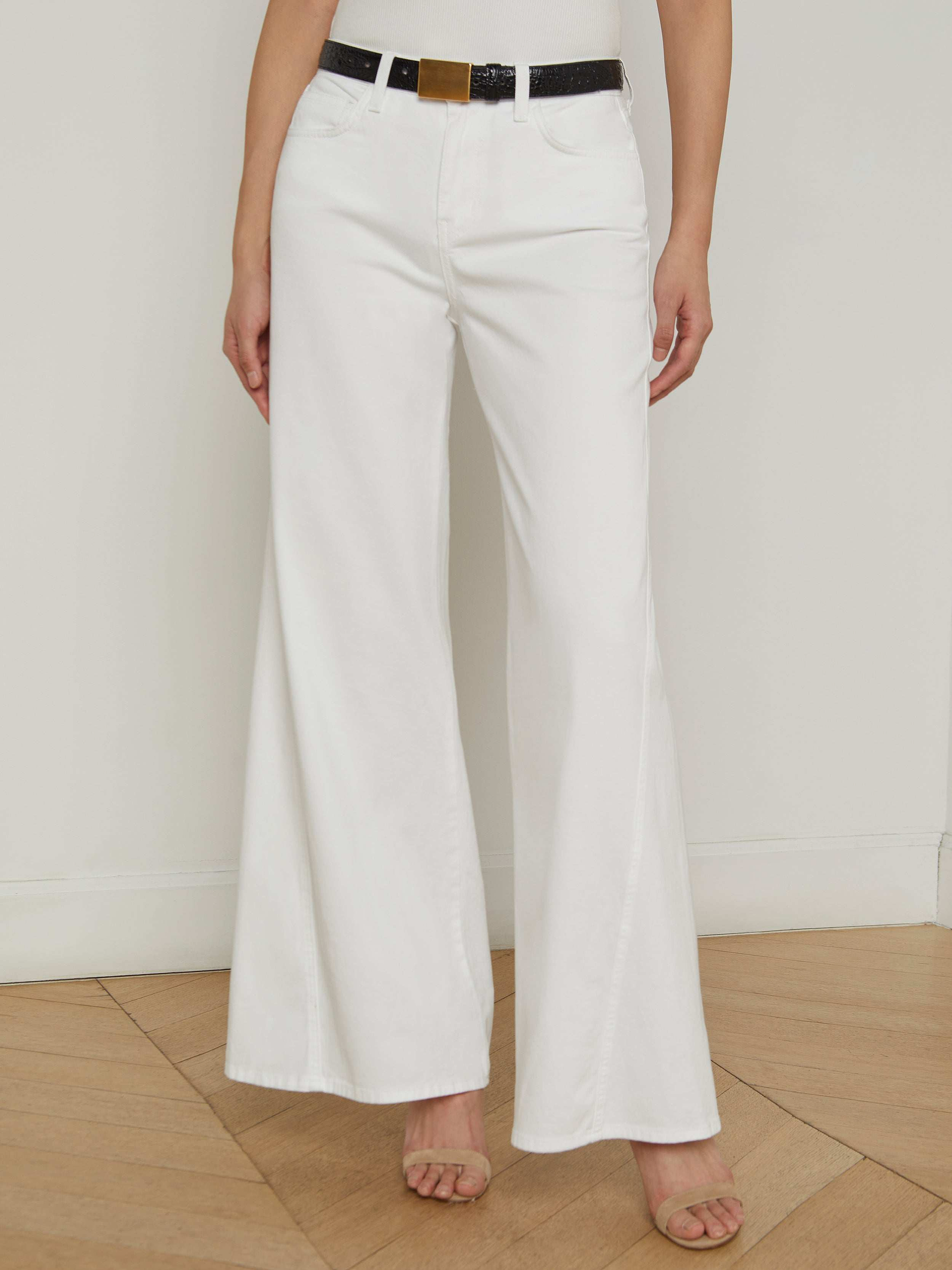 A person is shown from the waist down, wearing the Beau Wide-Leg Jean in white paired with a black belt and open-toed heels, standing against a plain light background. The upper body and face are not visible.