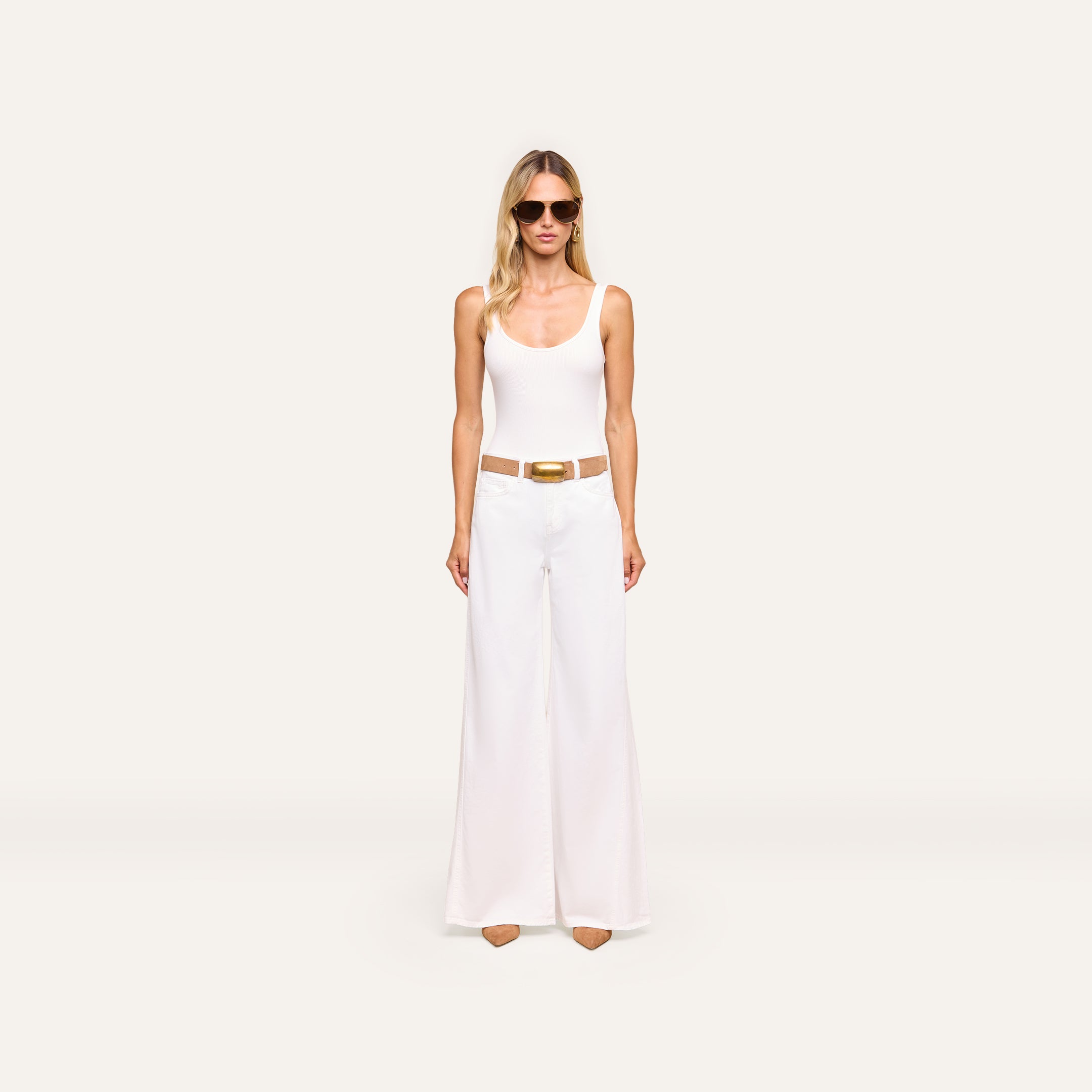 A woman in sunglasses, a white tank top, the Beau Wide-Leg Jean with a brown belt, and tan pointed shoes stands against an off-white background.