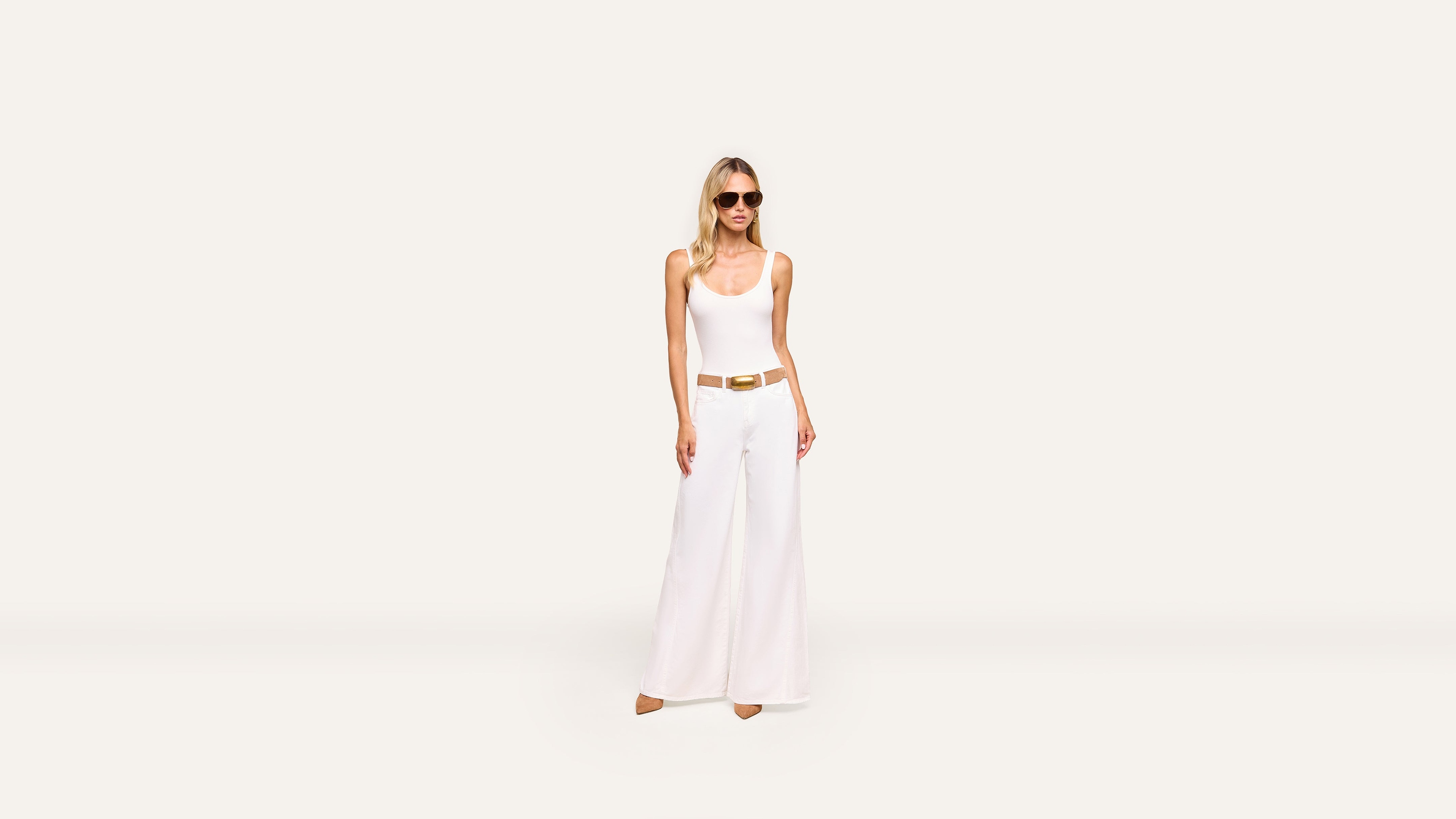 A woman wearing sunglasses, a white sleeveless top, and wide-leg white pants with a tan belt stands stylishly against a plain white background.