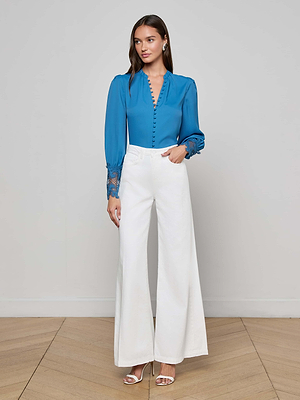 A woman stands indoors wearing a blue long-sleeve blouse with lace cuffs and L'AGENCE Quincy Wide-Leg Jeans in white, paired with white open-toe heels, pearl earrings, and her hair styled down.