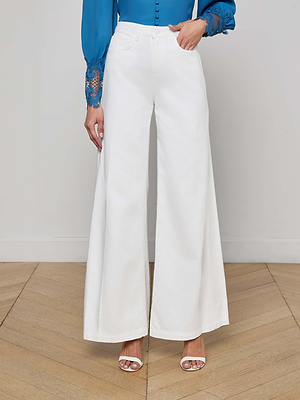 A person wears L'AGENCE Quincy Wide-Leg Jeans in white, a blue long-sleeve blouse with lace cuffs, and white open-toe heels while standing on a light wood floor against a plain wall.