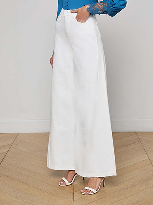 A person wearing L'AGENCE Quincy Wide-Leg Jean in white, paired with a blue lace-detailed long-sleeve blouse and white open-toe heels, stands on a wooden floor against a plain light background.