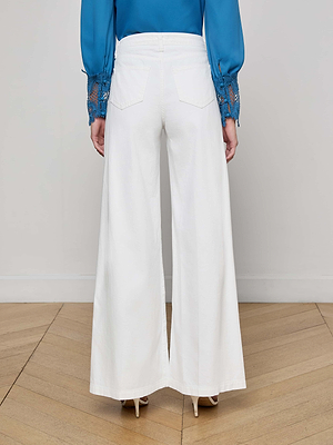 A person stands on a wooden floor facing away, wearing L'AGENCE Quincy Wide-Leg Jean in white, paired with a blue long-sleeve blouse featuring lace-detailed cuffs and beige high heels.