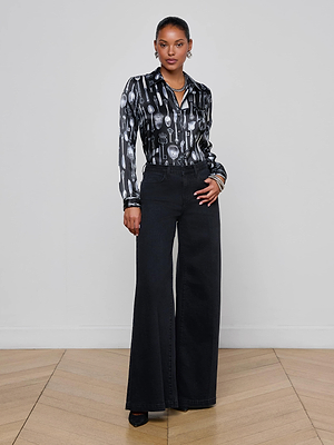 A woman poses confidently against a light gray wall, wearing the Quincy Wide-Leg Jean by L'AGENCE, paired with a black button-up shirt featuring a white spoon and fork pattern and black heels.