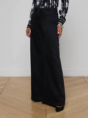Only the lower half of a person is visible, standing on a light wooden floor while wearing L'AGENCE Quincy Wide-Leg Jeans in black denim paired with a shiny, long-sleeved patterned blouse.