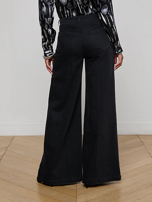 A person stands on a wooden floor, seen from behind, wearing L'AGENCE Quincy Wide-Leg Jeans in black denim and a long-sleeve shirt with a black and white utensil pattern.