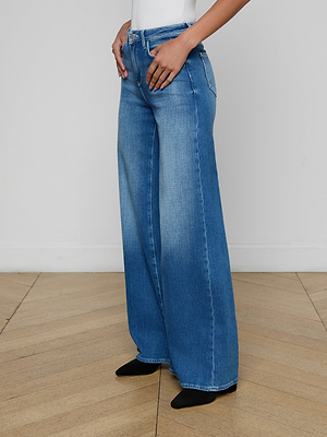Wearing the Quincy Wide-Leg Jean in aged indigo and a white top, a person stands on a wooden floor with hands on hips, only their lower half visible.
