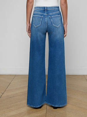 A person stands with their back to the camera wearing a white top and the Quincy Wide-Leg Jean, featuring a high rise, wide-leg fit in aged indigo with back pockets. The setting includes a light wood floor and plain white wall.