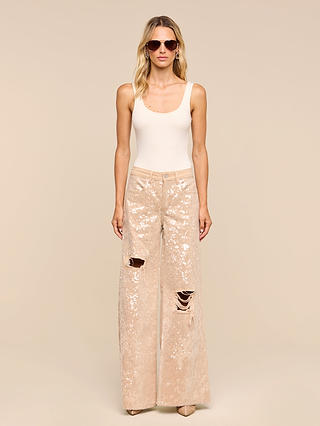 A woman stands facing forward against a plain beige background, wearing sunglasses, a sleeveless beige top, and the Quincy Embellished Wide-Leg Jean—high-rise sequined denim with ripped details.