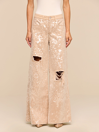 A person wearing the Quincy Embellished Wide-Leg Jean—high rise, wide-leg denim pants adorned with sequins and featuring two ripped knees—paired with a fitted white top, standing against a plain beige background.