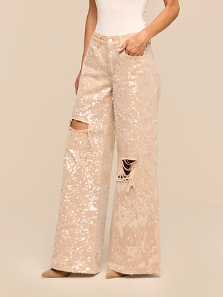 A person wearing the Quincy Embellished Wide-Leg Jean, featuring high-rise, wide-leg sequined denim with ripped details on both knees, paired with a white sleeveless top and pointed-toe shoes, stands against a plain beige background.