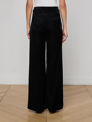 Person wearing Quincy Wide-Leg Corduroy Pant in black velvet corduroy with high rise and back pockets, standing on a wooden floor, facing away from the camera against a plain white wall.