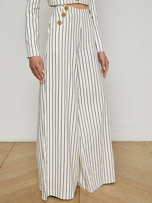A person wears the Kingston Striped Wide-Leg Pant by L'AGENCE Sale, featuring vertical black stripes and gold buttons, paired with a matching cropped jacket. They stand on a wooden floor against a plain white wall.