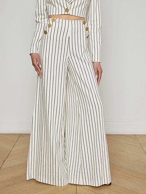A person wearing the L'AGENCE Sale Kingston Striped Wide-Leg Pant, featuring gold buttons near the waist, stands on a wooden floor against a plain white wall.