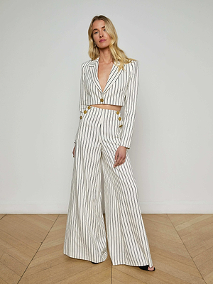 A woman with long blonde hair poses indoors against a plain wall and wooden floor, wearing L'AGENCE Sale's Kingston Striped Wide-Leg Pant in white with vertical black stripes, paired with a matching cropped blazer featuring gold buttons.