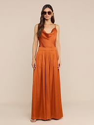 A woman wearing sunglasses and an orange sleeveless cowl-neck maxi dress stands against a plain white background, complemented by the fashionable Haylo Pant in burnt orange.
