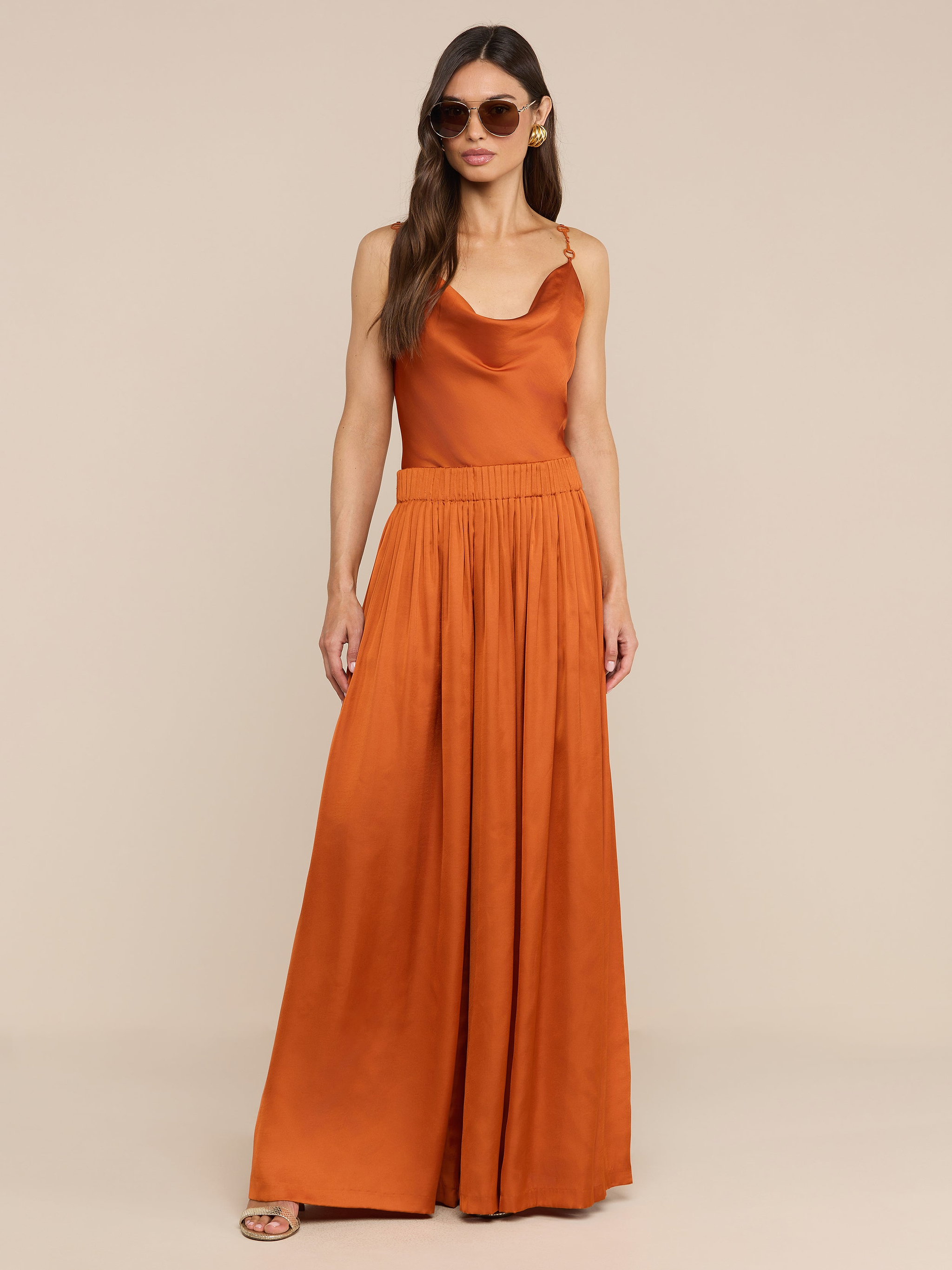 A woman wearing sunglasses and the Haylo Pant in burnt orange with a satin wide leg design stands against a plain white background.