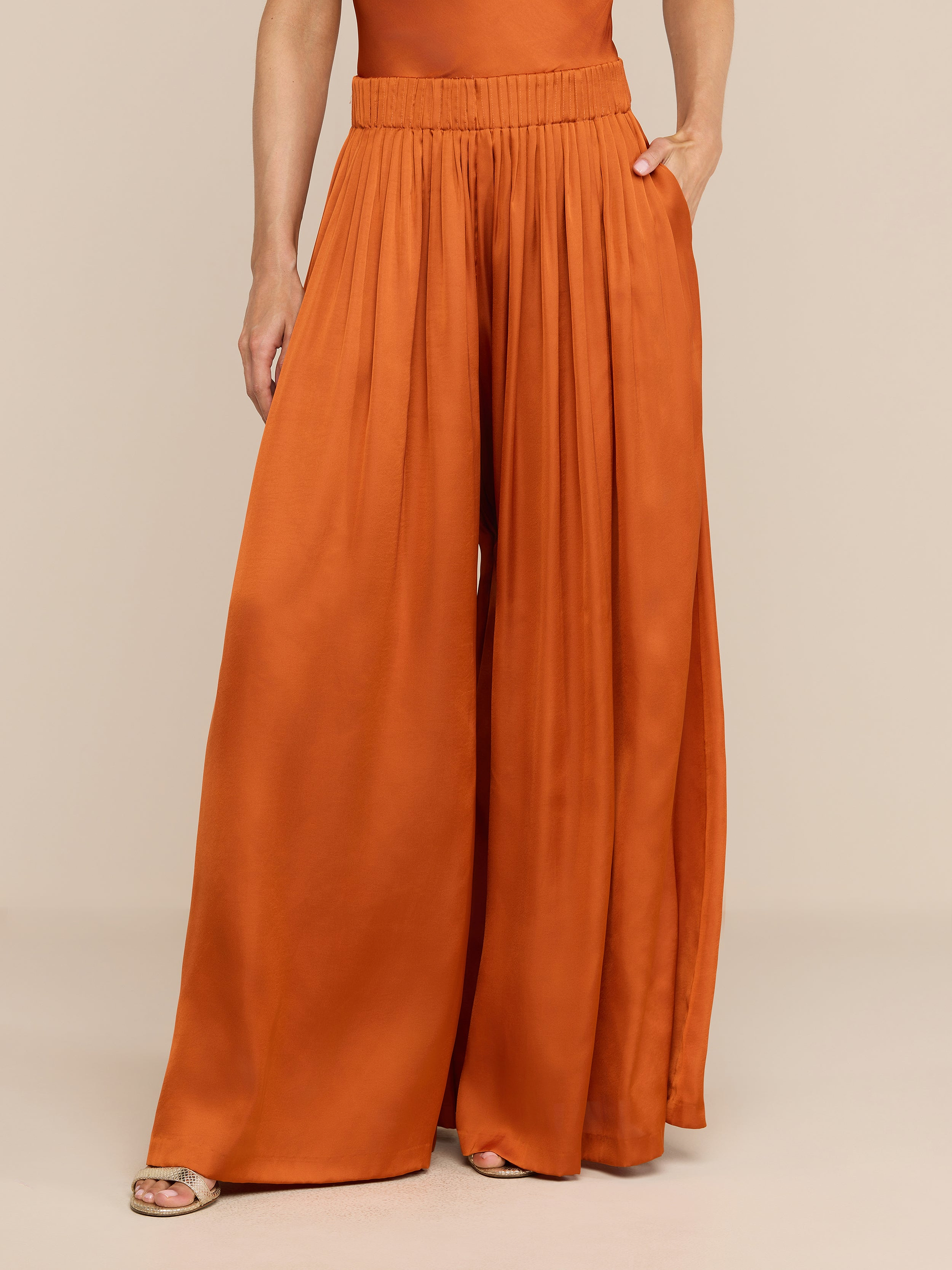 Person wearing the Haylo Pant in burnt orange—a high-waisted, satin wide-leg style with pockets—and open-toed sandals, standing against a plain white background. Upper torso and head are out of frame.
