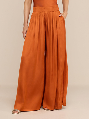 Model showcases the Haylo Pant in burnt orange—a high-waist satin wide leg style with a gathered waistband and side pockets. Only the lower torso and legs are visible; background is neutral.