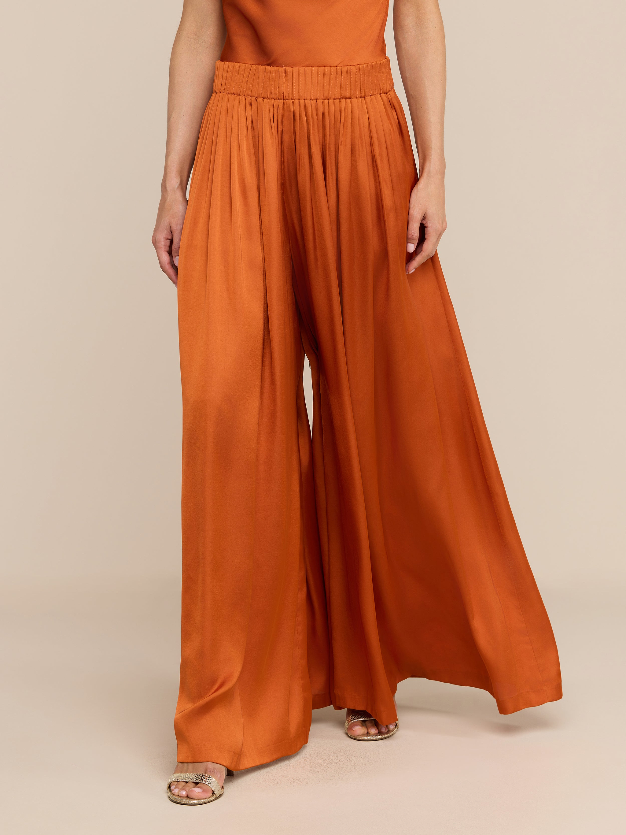 Wearing the Haylo Pant—high-waisted satin wide leg pants in burnt orange with an elastic waistband—paired with open-toe sandals, a person stands against a plain white background.