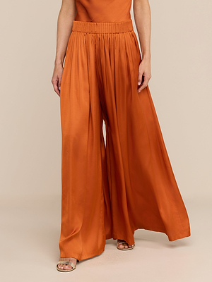 A person wearing Haylo Pant—satin, wide-leg, high-waisted in burnt orange with a gathered waistband—paired with open-toe sandals and an orange top, stands against a beige background.