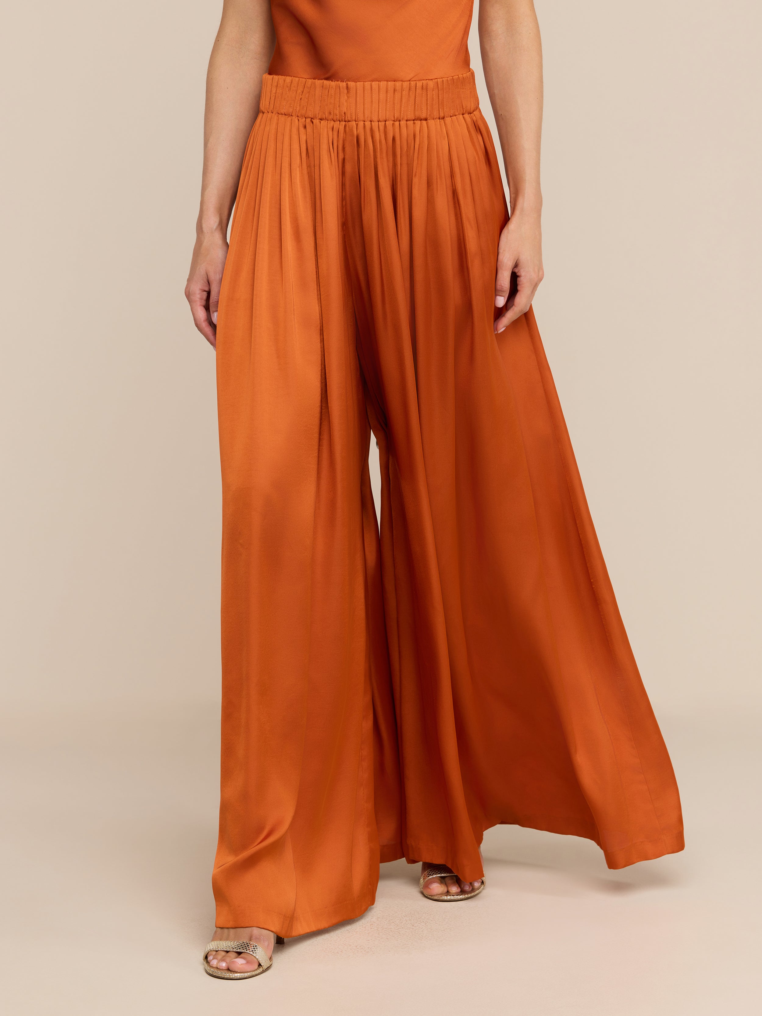 Wearing the Haylo Pant—high-waisted satin wide leg pants in burnt orange with an elastic waistband—paired with open-toe sandals, a person stands against a plain white background.