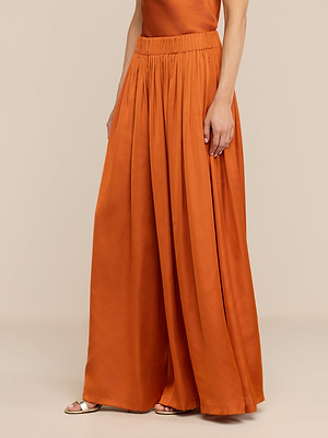 A person wearing the Haylo Pant in high waist, burnt orange style with a matching sleeveless top and open-toe sandals, standing against a plain beige background.