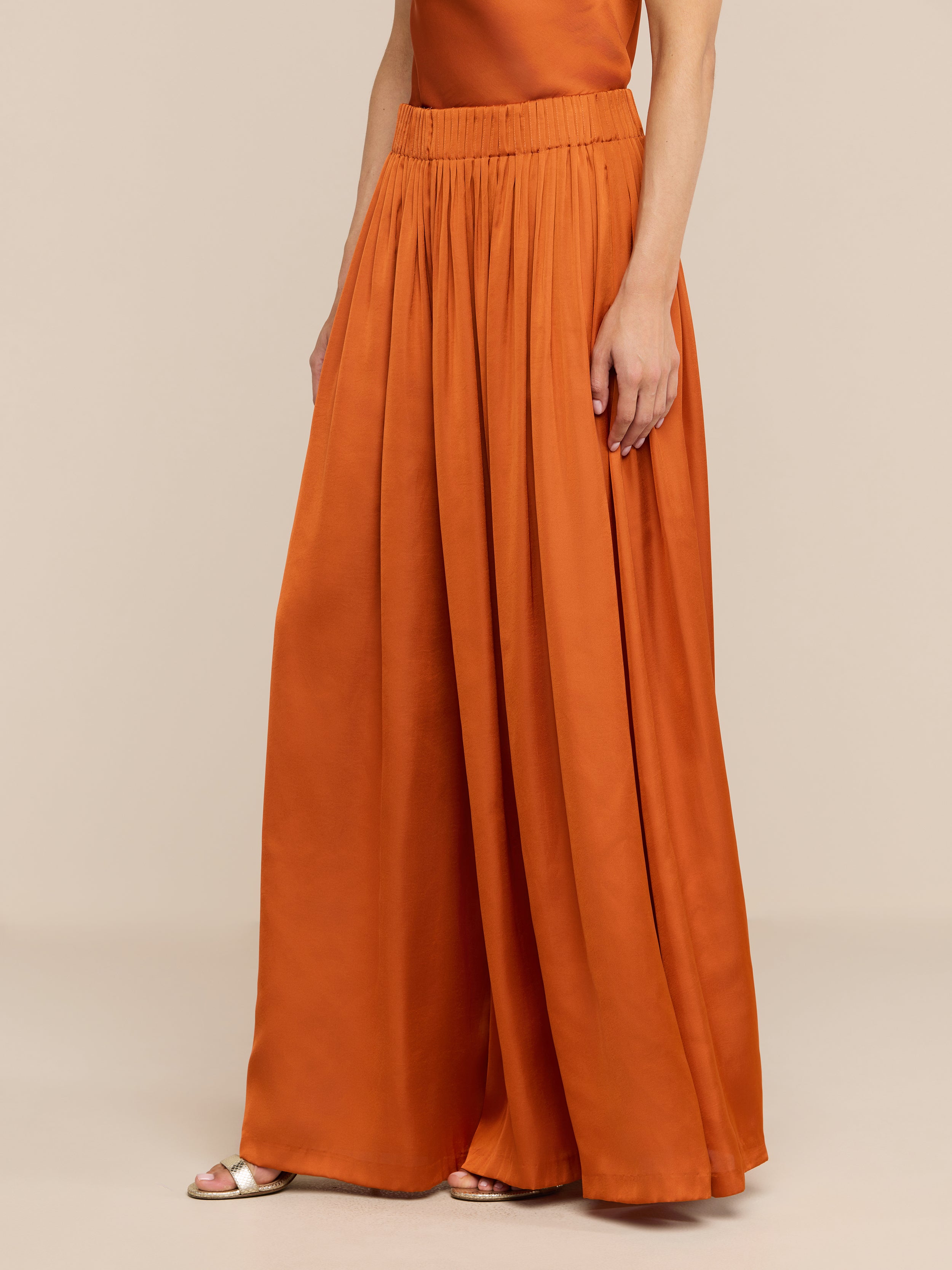 A woman wears the Haylo Pant, a pair of high waist burnt orange satin wide-leg pants, styled with a matching sleeveless top and sandals, standing against a plain white background.