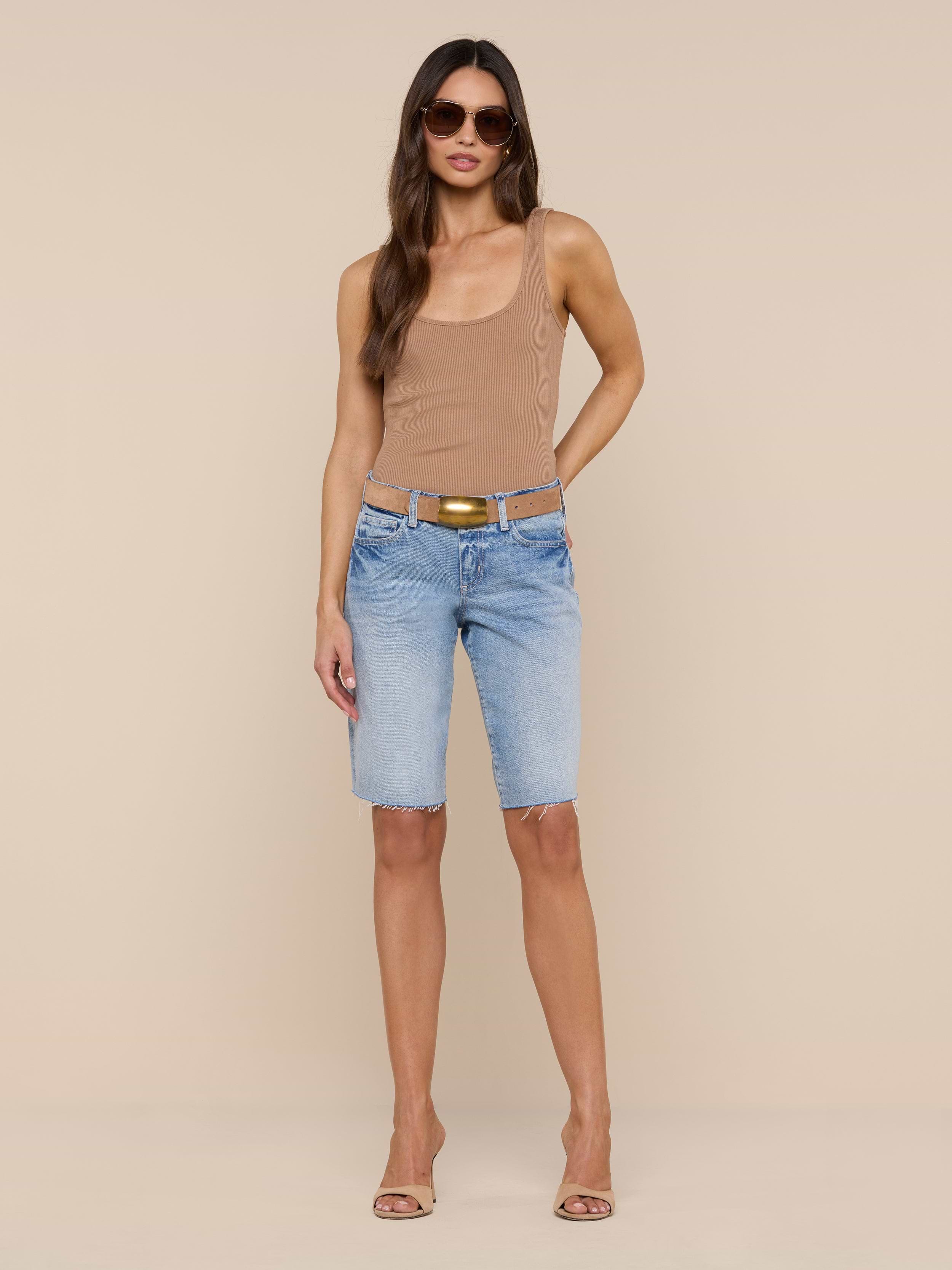 A woman stands against a plain white background wearing sunglasses, a tan sleeveless top, heeled sandals, and the Elise Jean Bermuda Short—light blue low-rise shorts crafted from all-cotton denim with a relaxed fit.