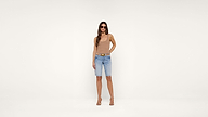 A woman wearing sunglasses, a beige tank top, blue knee-length denim shorts, and beige heels stands against a plain white background.
