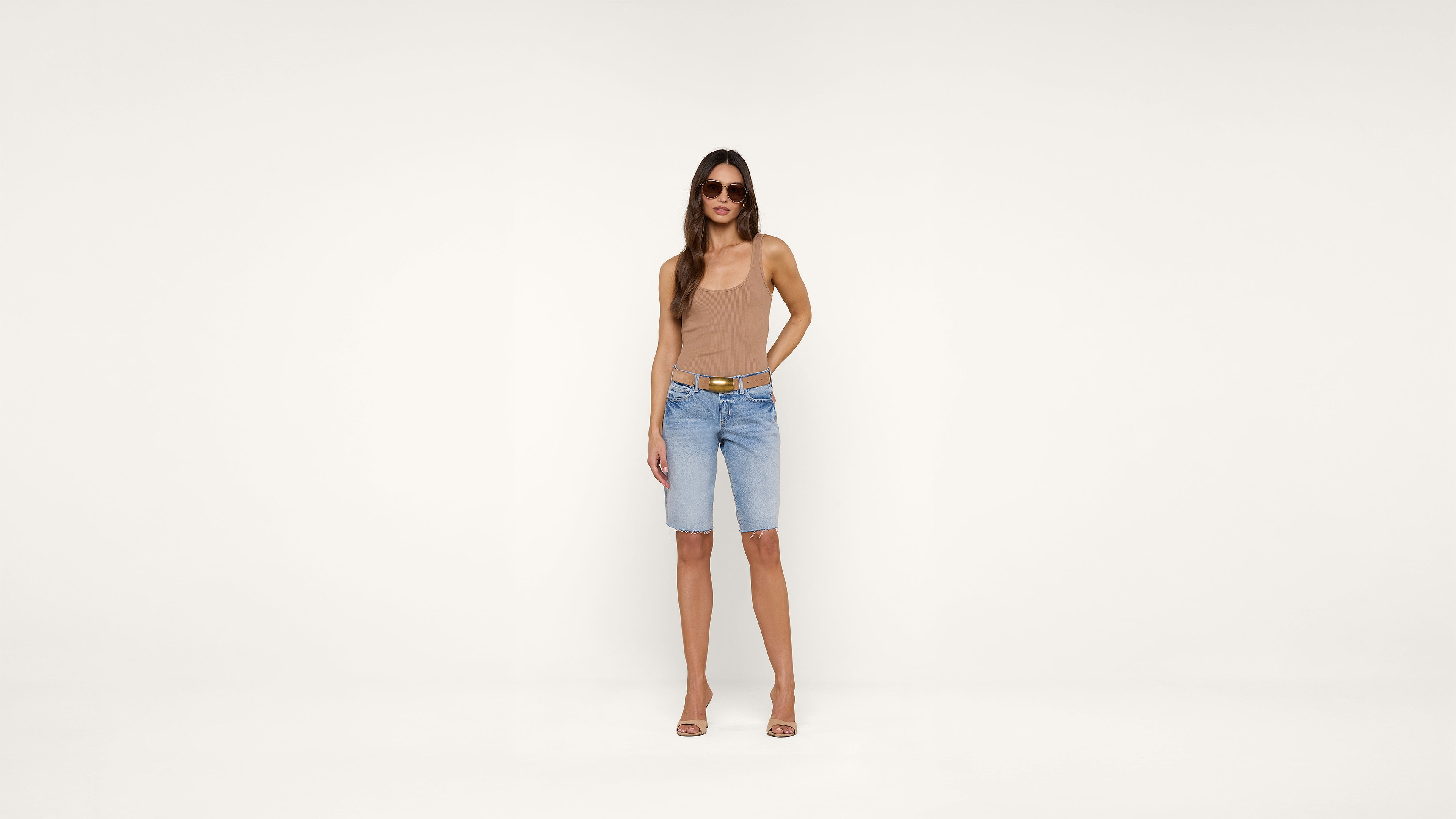 A woman wearing sunglasses, a beige tank top, blue knee-length denim shorts, and beige heels stands against a plain white background.