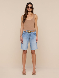 A woman stands against a plain white background wearing sunglasses, a tan tank top, and the Elise Jean Bermuda Short—light blue low-rise shorts with a relaxed fit crafted from all-cotton denim. She completes her look with heeled sandals.