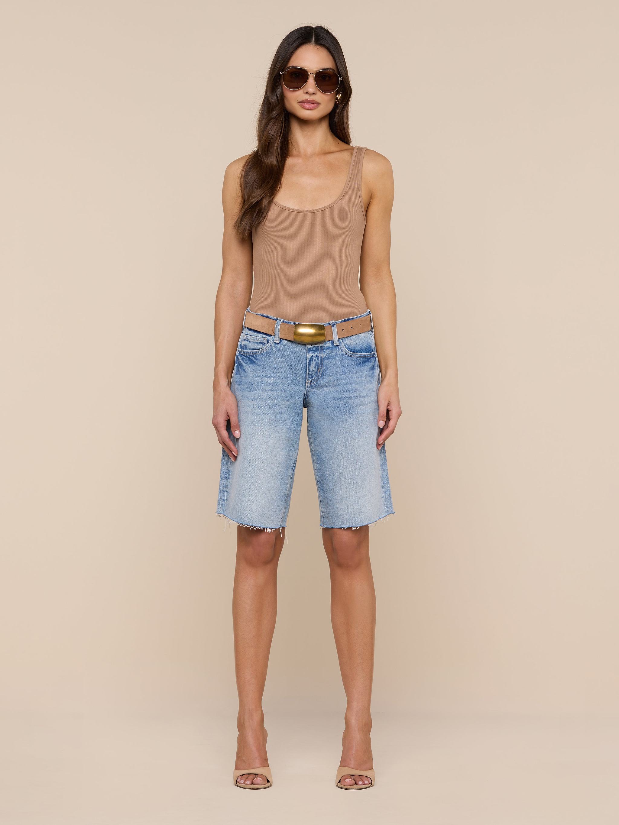 A woman stands against a plain white background wearing sunglasses, a tan tank top, and the Elise Jean Bermuda Short—light blue low-rise shorts with a relaxed fit crafted from all-cotton denim. She completes her look with heeled sandals.