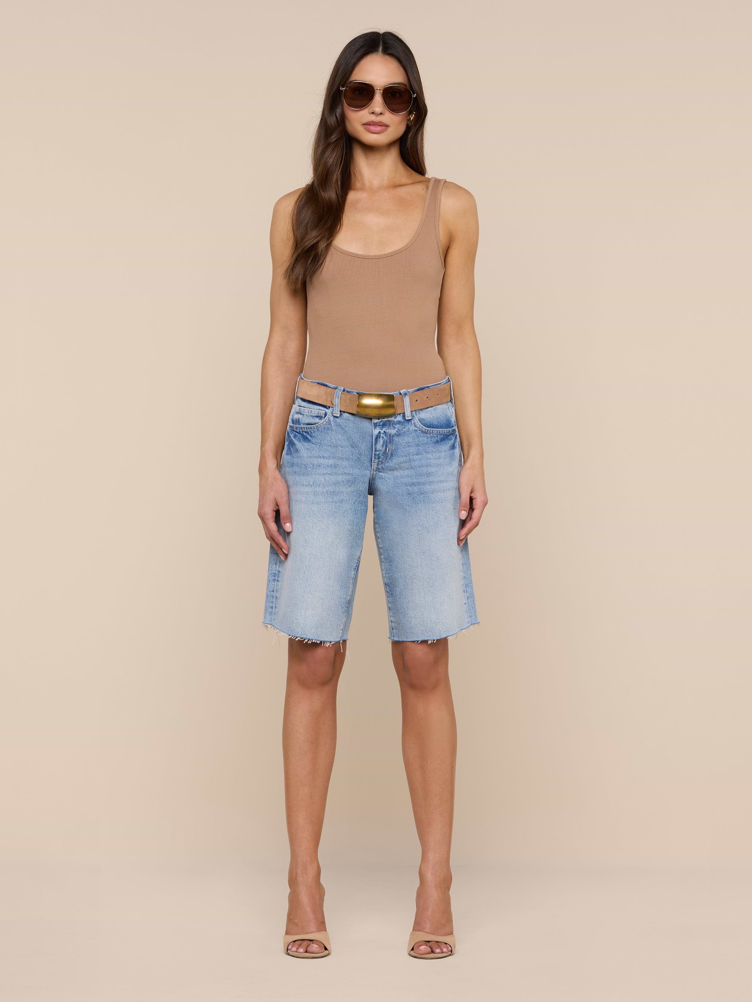A woman stands against a plain white background wearing sunglasses, a tan tank top, and the Elise Jean Bermuda Short—light blue low-rise shorts with a relaxed fit crafted from all-cotton denim. She completes her look with heeled sandals.