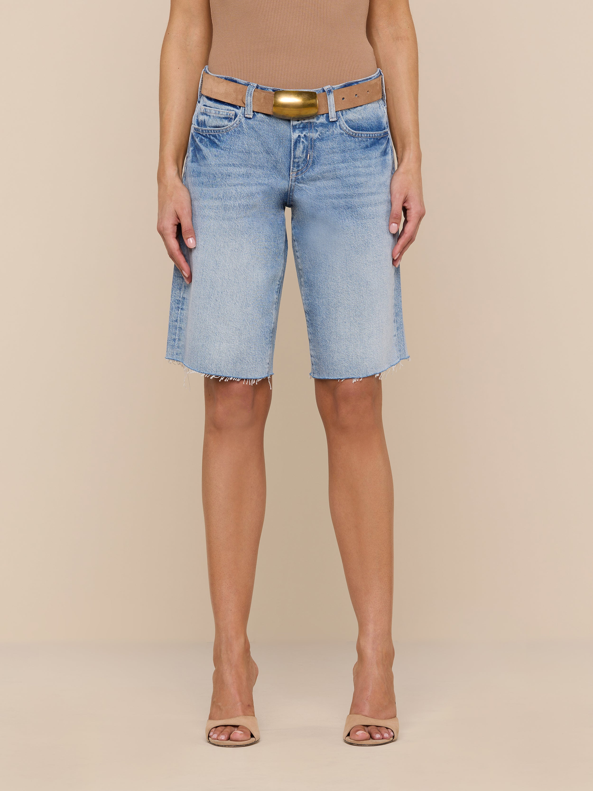 A person wearing a tan sleeveless top, a tan belt, open-toe heels, and the Elise Jean Bermuda Short—a relaxed fit, light blue, knee-length all-cotton denim short with a raw hem—stands against a plain white background.
