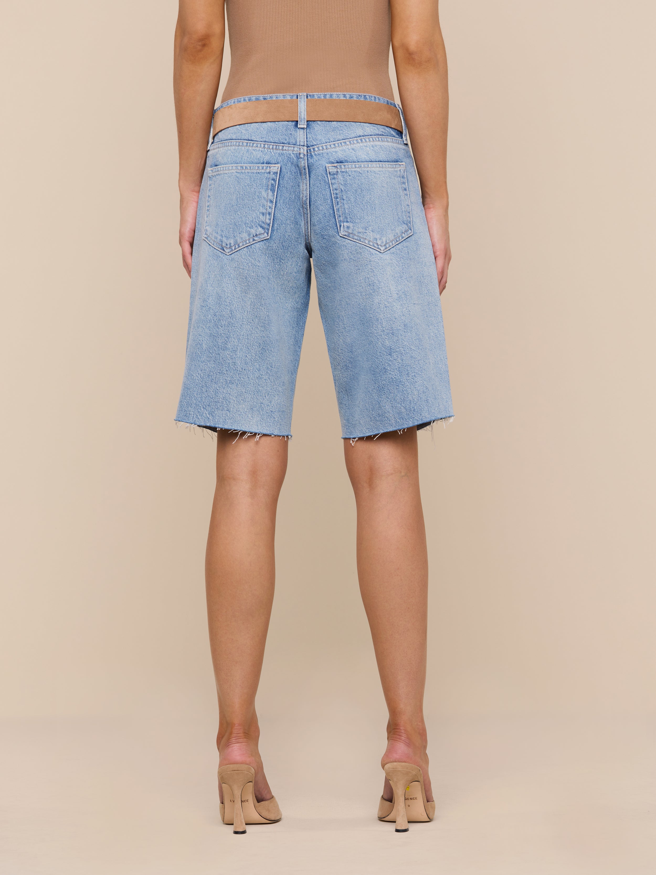 A person is shown from the back against a plain white background, wearing a tan top, nude high heels, and the Elise Jean Bermuda Short—relaxed fit, all-cotton denim knee-length shorts in light blue.