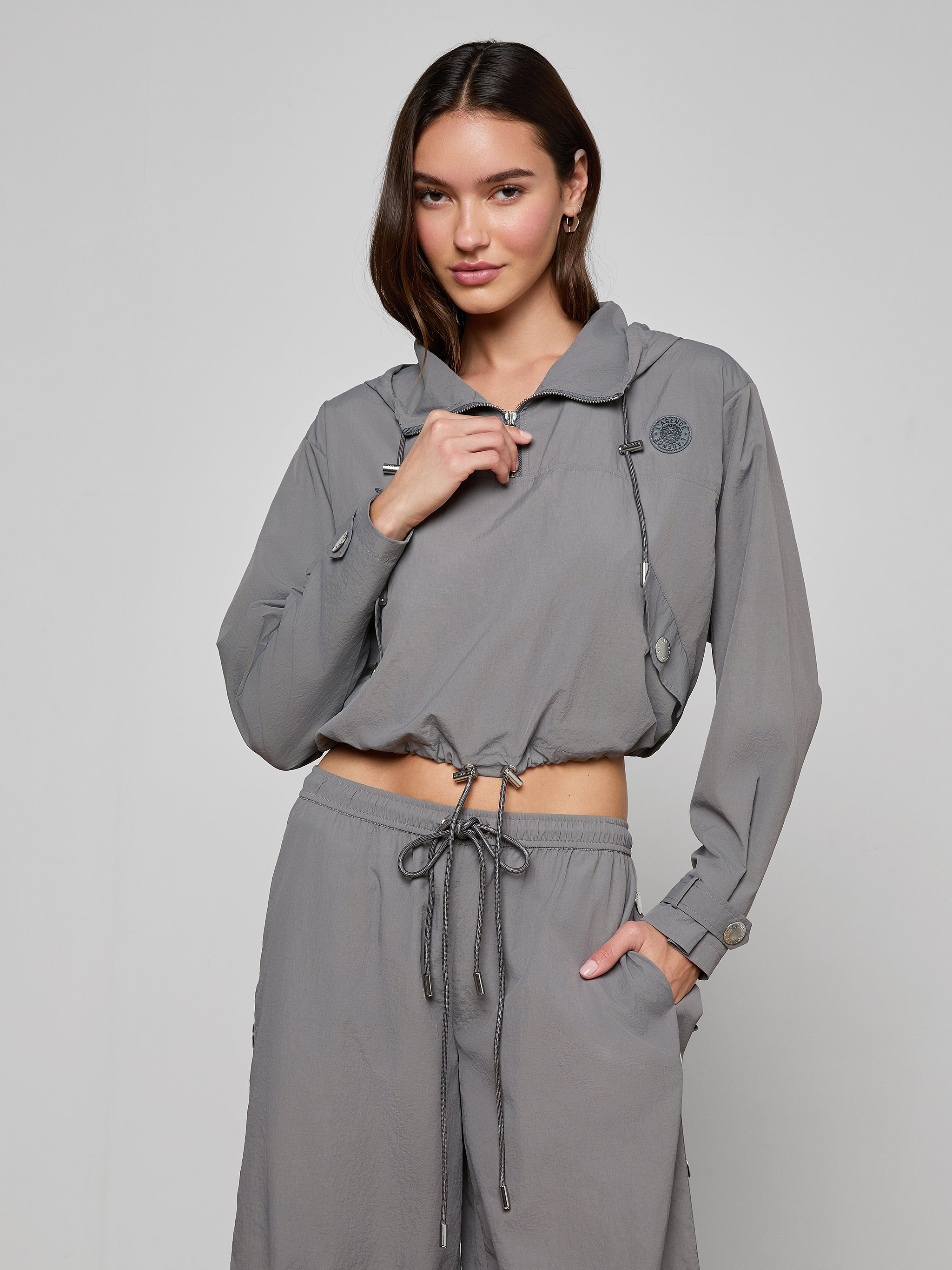 A woman stands against a plain light background wearing the Bronwyn Cropped Windbreaker in light gray, featuring long sleeves, a hood, and a cropped silhouette, paired with matching drawstring pants. She holds the zipper near her neck with one hand.