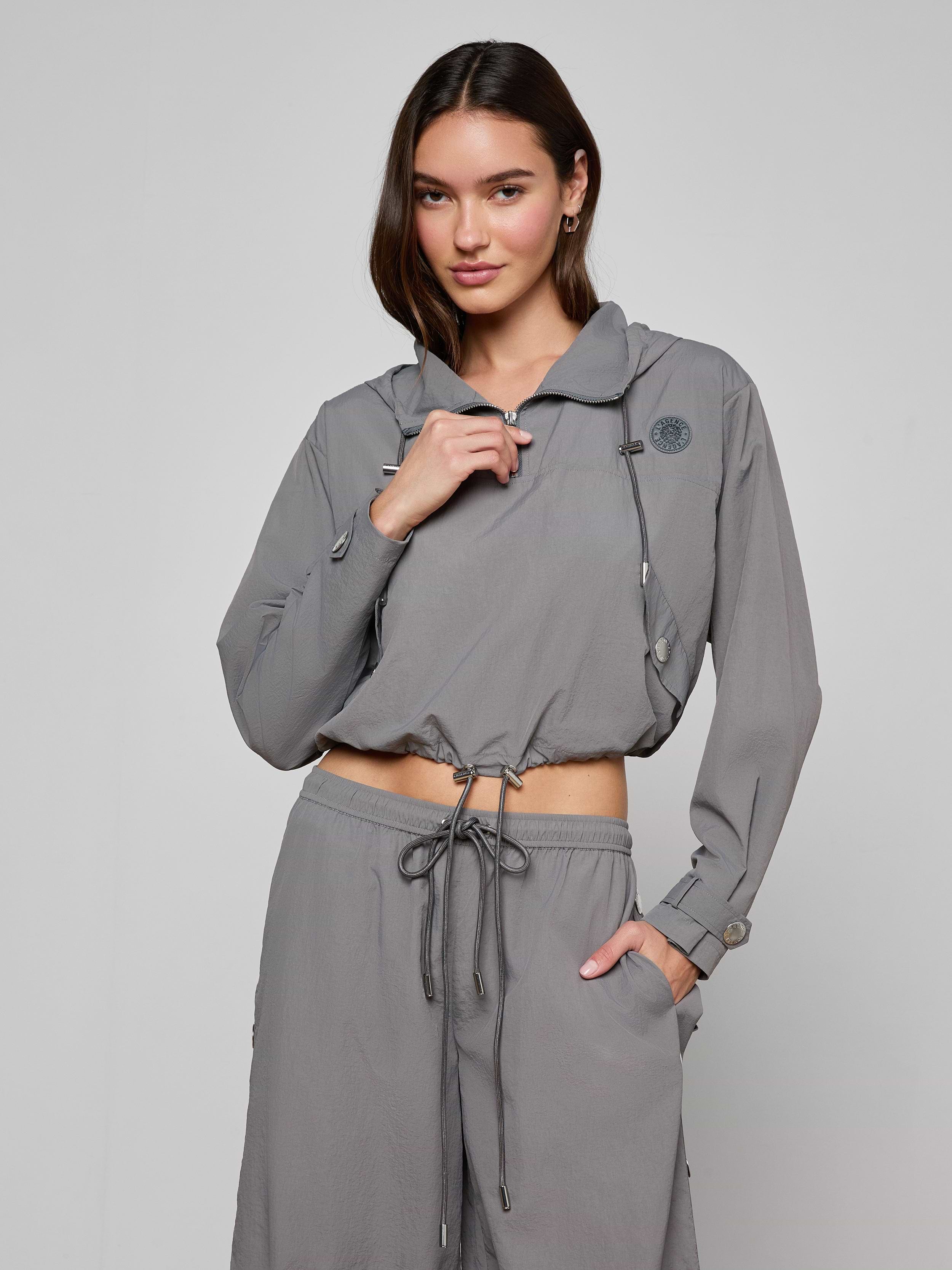 A woman stands against a plain light background wearing the Bronwyn Cropped Windbreaker in light gray, featuring long sleeves, a hood, and a cropped silhouette, paired with matching drawstring pants. She holds the zipper near her neck with one hand.