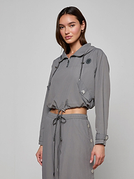 A woman stands against a plain light background wearing the Bronwyn Cropped Windbreaker—a gray, long-sleeved, collared jacket with drawstrings—paired with matching high-waisted pants.