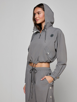 Wearing the Bronwyn Cropped Windbreaker and matching drawstring pants, a woman stands with one hand in her pocket, looking to the side. The water-repellent jacket has silver buttons and a front zipper.