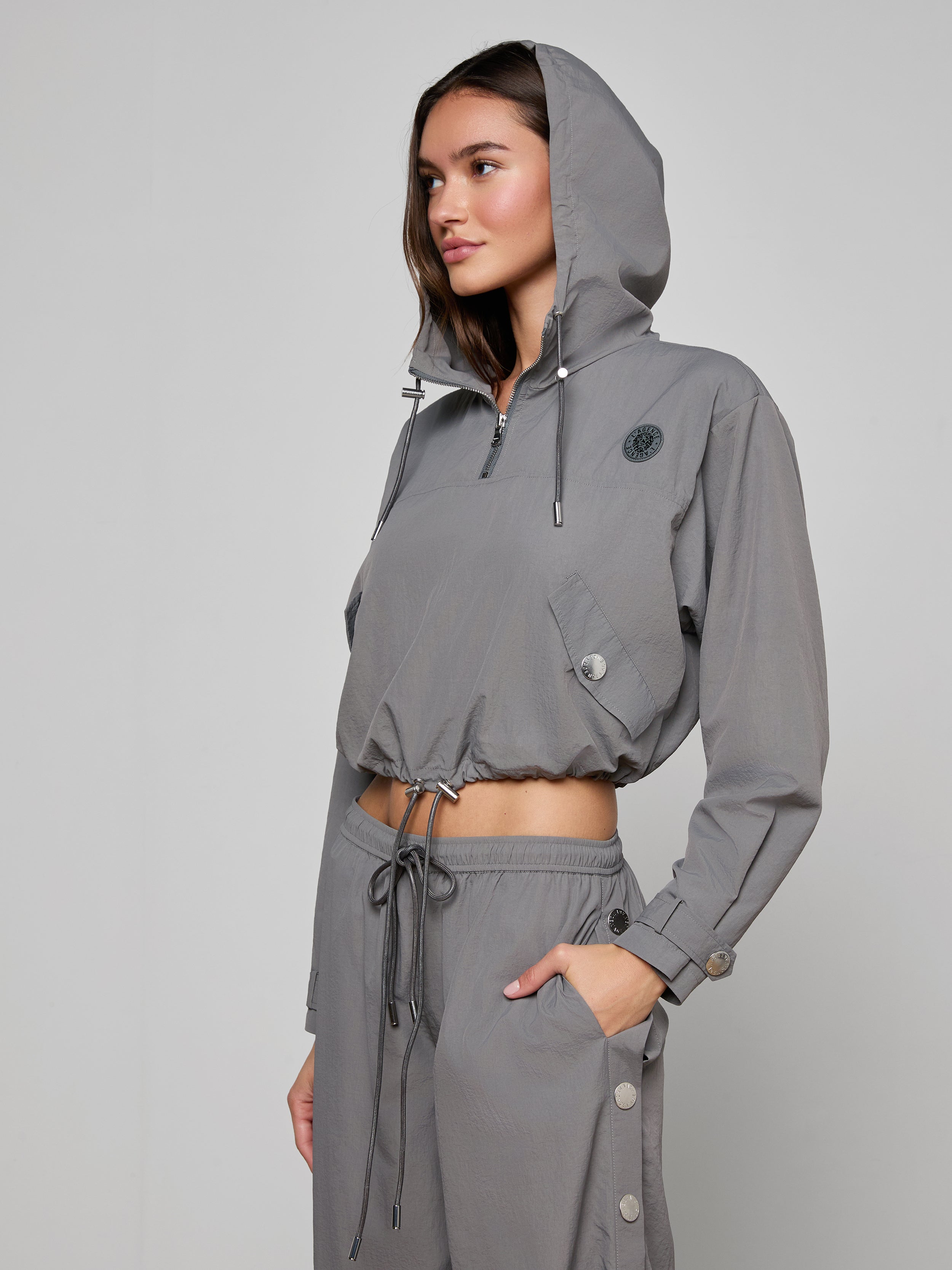 A woman wearing the Bronwyn Cropped Windbreaker and matching pants stands against a plain light background, looking to the side with one hand in her pocket.