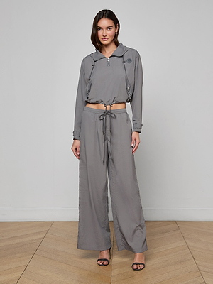 A woman with shoulder-length dark hair stands indoors on a wooden floor, wearing a gray cropped hooded jacket and matching Valkyrie Snap Pant, styled with black heeled sandals.