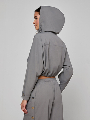 A woman stands sideways against a plain light background, wearing the Bronwyn Cropped Windbreaker in light gray with the hood up and her left hand on her hip, paired with matching high-waisted pants featuring button accents.