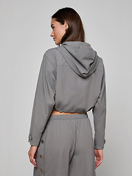 A woman stands facing away, wearing the Bronwyn Cropped Windbreaker in gray, paired with matching pants featuring button details along the sides. The outfit showcases a relaxed, casual style with a cropped silhouette against a plain light background.