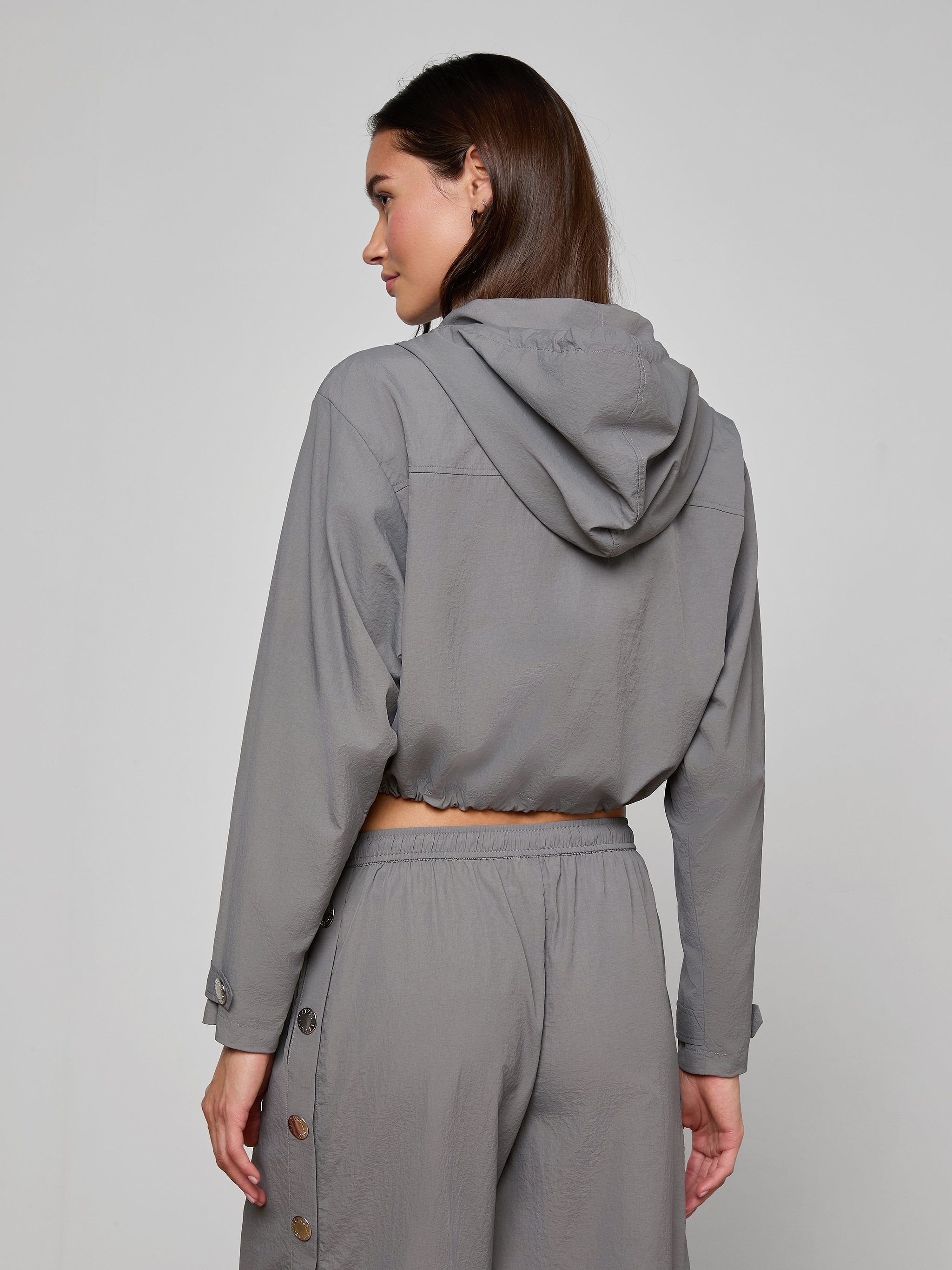 A woman stands facing away, wearing the Bronwyn Cropped Windbreaker in gray, paired with matching pants featuring button details along the sides. The outfit showcases a relaxed, casual style with a cropped silhouette against a plain light background.