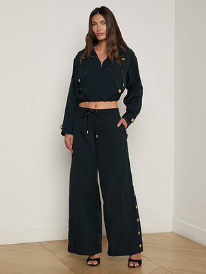 A woman with long brown hair and a neutral expression stands indoors, wearing loose-fitting black Valkyrie Snap Pants with wide legs, a cropped long-sleeve top with gold buttons, and black heeled sandals.