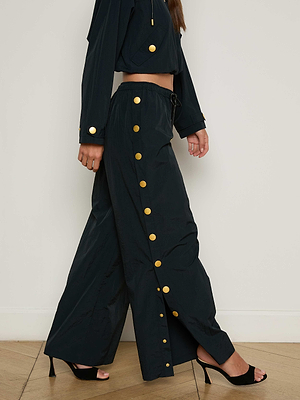 A person wearing black Valkyrie Snap Pant with large gold side buttons, a matching black top with gold button details, and black open-toe heels stands on a wooden floor against a plain wall.