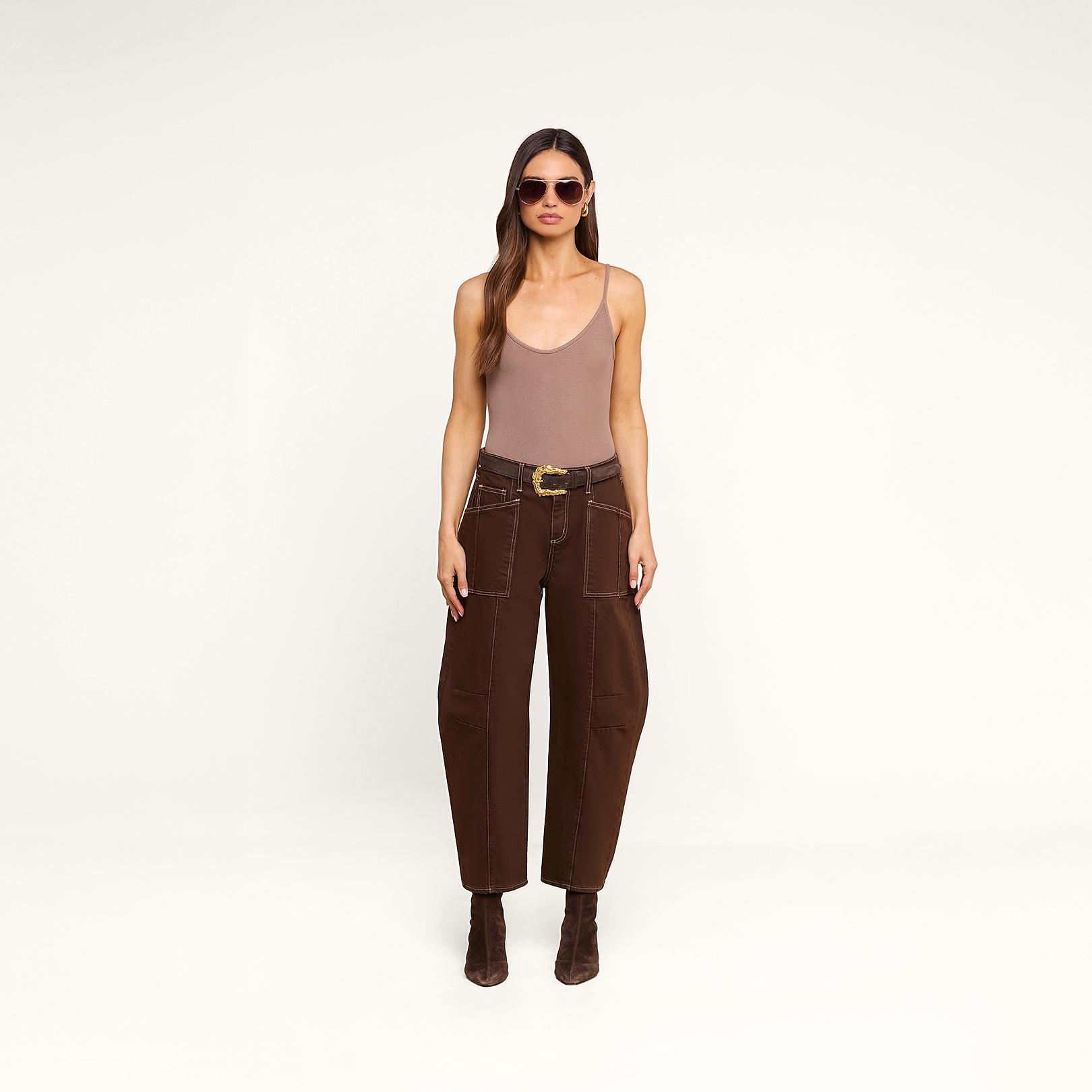 A woman with long brown hair wears sunglasses, a sleeveless taupe bodysuit, the Cody Cropped Barrel-Leg Jean in high-rise dark brown non-stretch denim with a wide belt, and brown boots, standing against a beige background.
