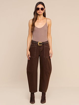 A woman with long brown hair wears sunglasses, a sleeveless taupe bodysuit, the Cody Cropped Barrel-Leg Jean in high-rise dark brown non-stretch denim with a wide belt, and brown boots, standing against a beige background.