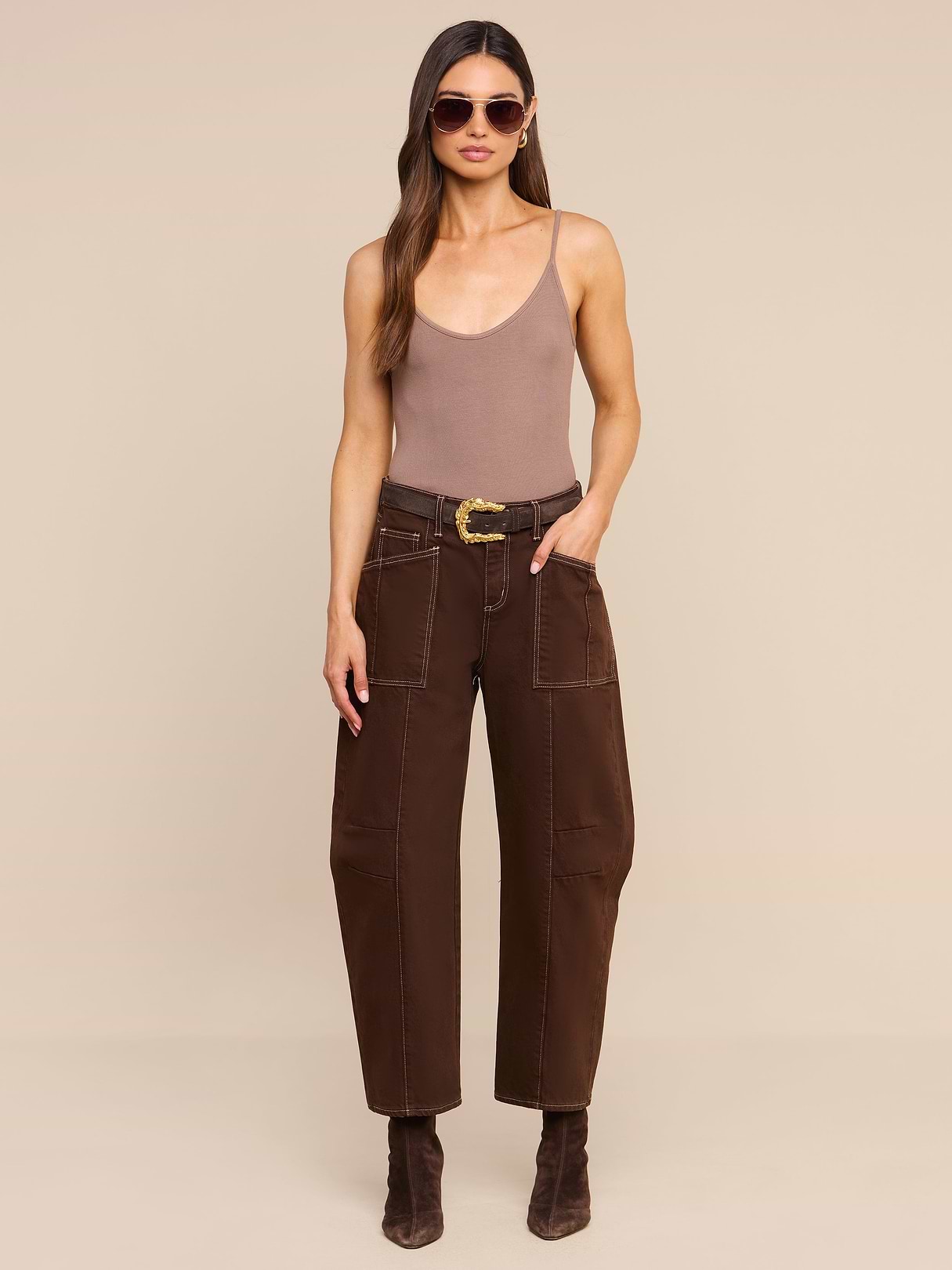 Woman wearing sunglasses, a brown tank top, the Cody Cropped Barrel-Leg Jean in high-rise dark brown non-stretch denim with a belt, and brown boots stands against a beige background.